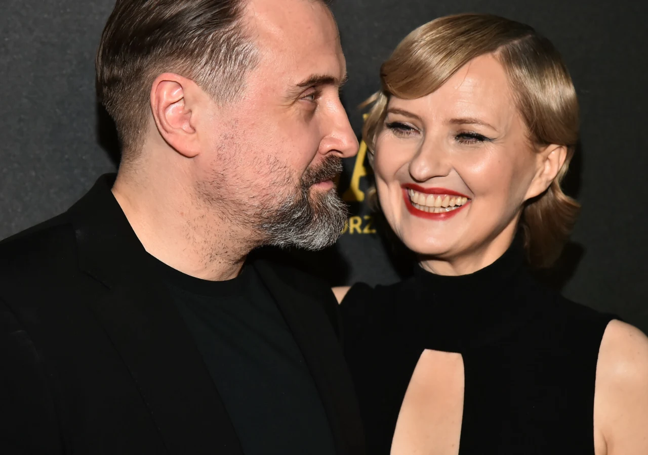 Marek Modzelewski, Izabela Kuna Para elegancko ubranych osób, mężczyzna z zarostem spogląda na uśmiechniętą kobietę o jasnych włosach, oboje wyglądają na szczęśliwych, tło w ciemnych kolorach z częściowo widocznym napisem.