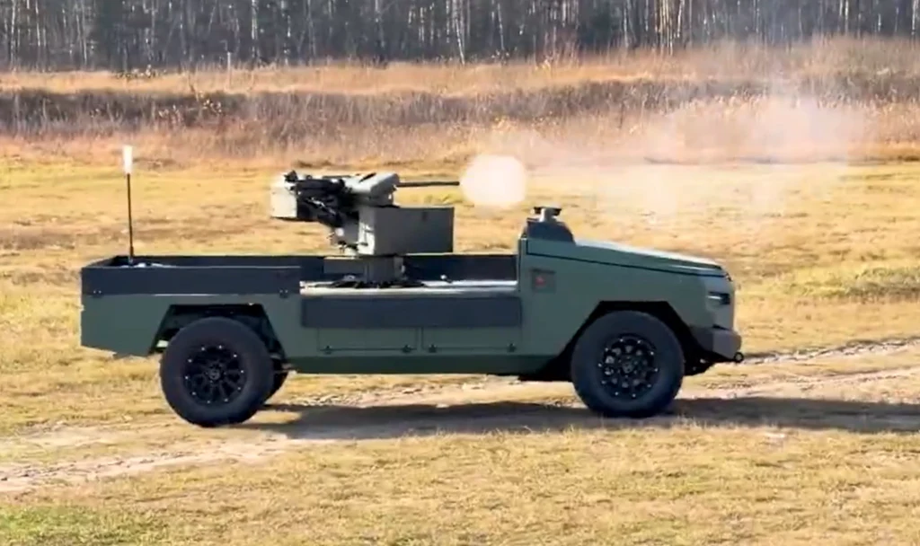 Wojskowy pojazd bezzałogowy z zamontowanym systemem uzbrojenia, który wykonuje testowy strzał na otwartym terenie poligonu, na tle wysuszonej trawy i ściany lasu.