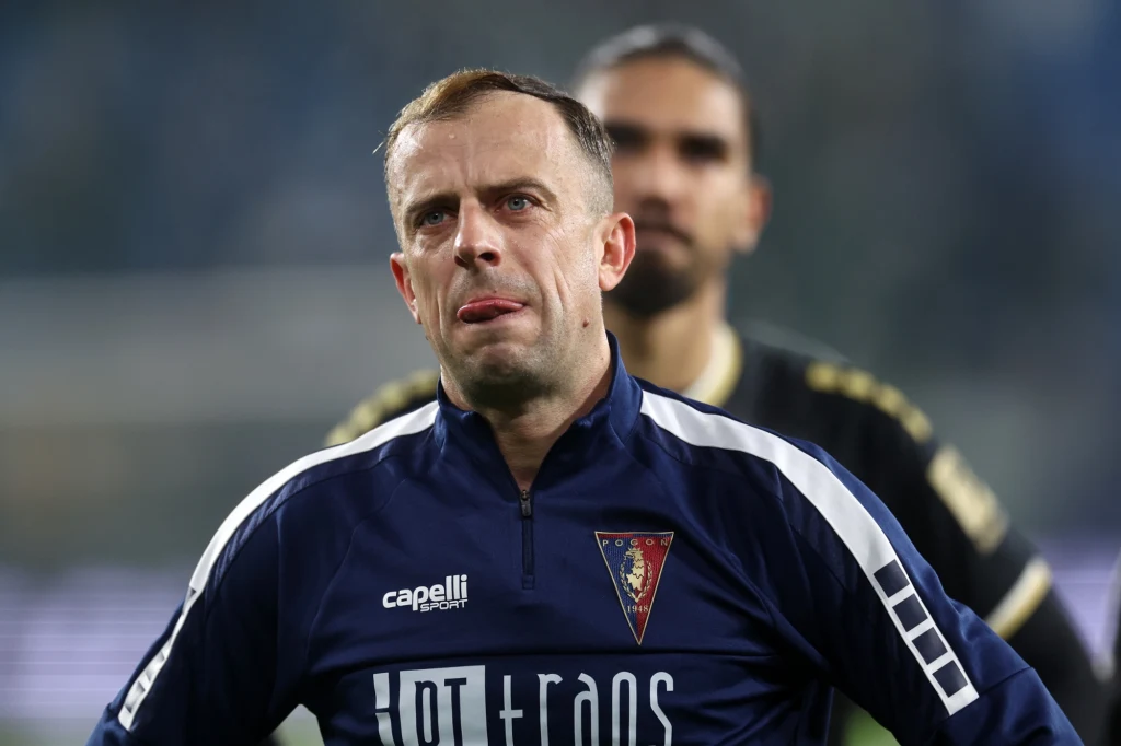 Kamil Grosicki Piłkarz w granatowej koszulce z logotypem klubu sportowego stoi na pierwszym planie, za nim widoczna jest druga postać w sportowym stroju. Tło rozmyte, akcent na wyraz twarzy sportowca.