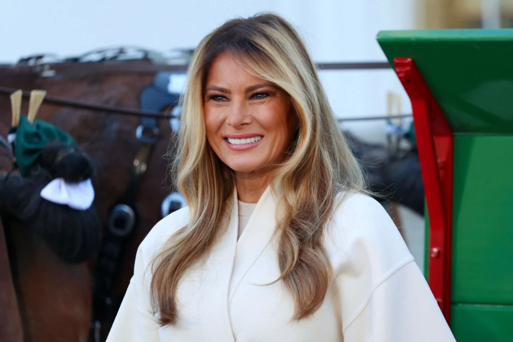 Melania Trump odebrała świąteczne drzewko, które stanie w Białym Domu Kobieta o jasnych, długich włosach uśmiecha się stojąc na tle konia i fragmentu bryczki. Ubrana jest w jasny płaszcz, ma naturalny makijaż i wygląda pogodnie.