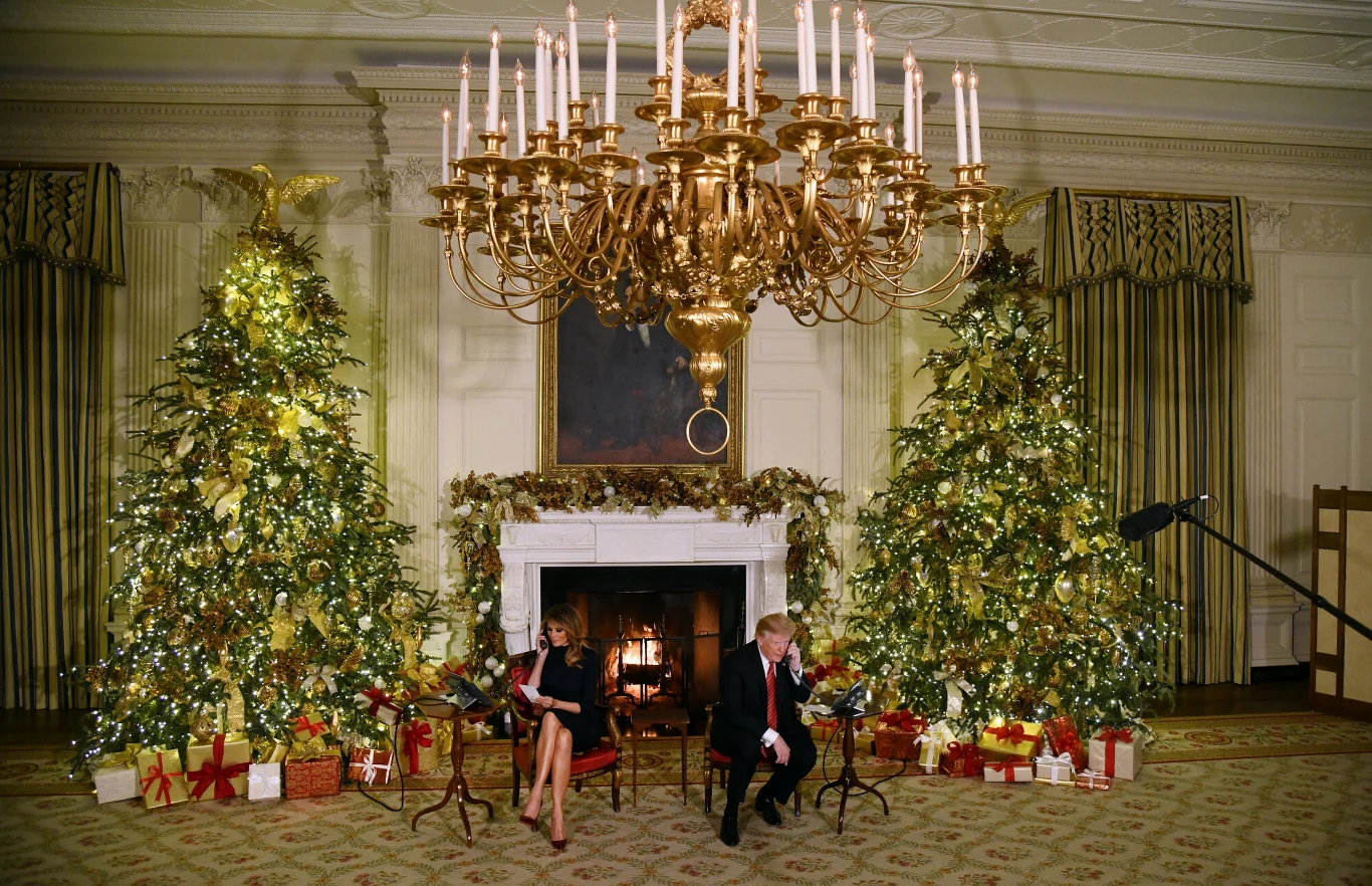 Dwie osoby siedzące przed kominkiem w eleganckim, bogato udekorowanym pokoju, otoczone dużymi choinkami przystrojonymi świątecznymi ozdobami i prezentami, całość oświetlona dużym, złotym żyrandolem.