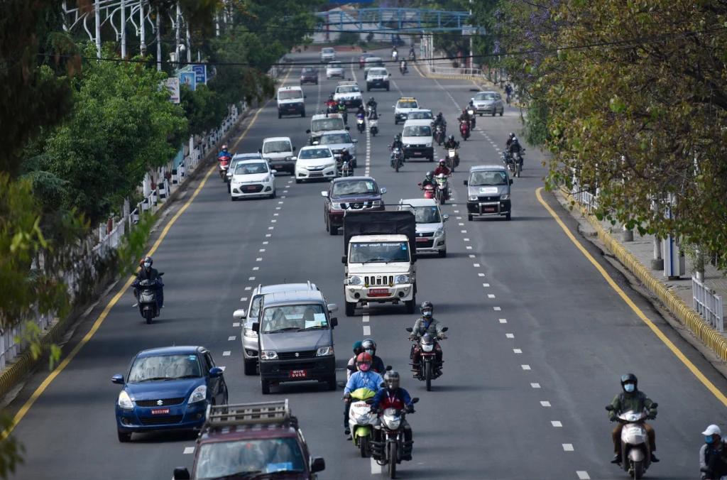 Główny element stanowią samochody oraz motocykle poruszające się po szerokiej miejskiej ulicy otoczonej drzewami i ogrodzeniem. Kierowcy jadą w obu kierunkach, a ruch przebiega płynnie w środku dnia.