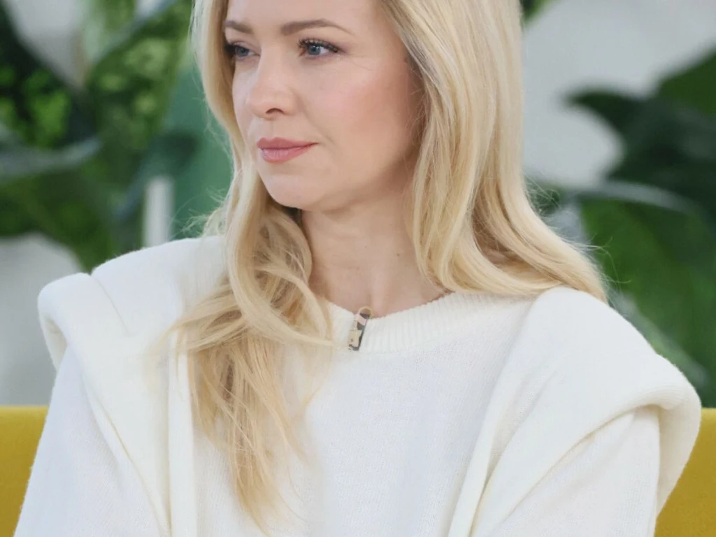 Blondynka o jasnej karnacji w białym swetrze siedzi na żółtym fotelu, w tle zielone liście roślin.