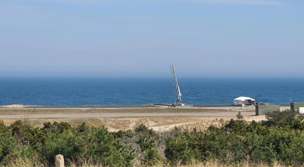 Udany start polskiej rakiety. Wystartowała z poligonu w Ustce. Stalowa konstrukcja przypominająca rampę startową lub urządzenie do wystrzeliwania rakiet ustawiona nad wybrzeżem morza. W tle rozciąga się szeroka tafla wody, a w pierwszym planie znajduje się niska zieleń. Obok konstrukcji widoczny biały namiot lub m...