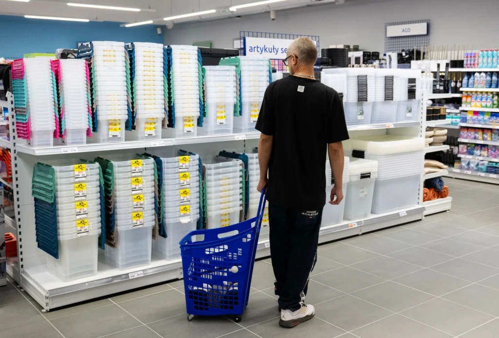 Mężczyzna z niebieskim koszykiem na kółkach spaceruje po sklepie, oglądając plastikowe pudełka i pojemniki ustawione na półkach. W tle widoczny dobrze oświetlony dział z artykułami gospodarstwa domowego.