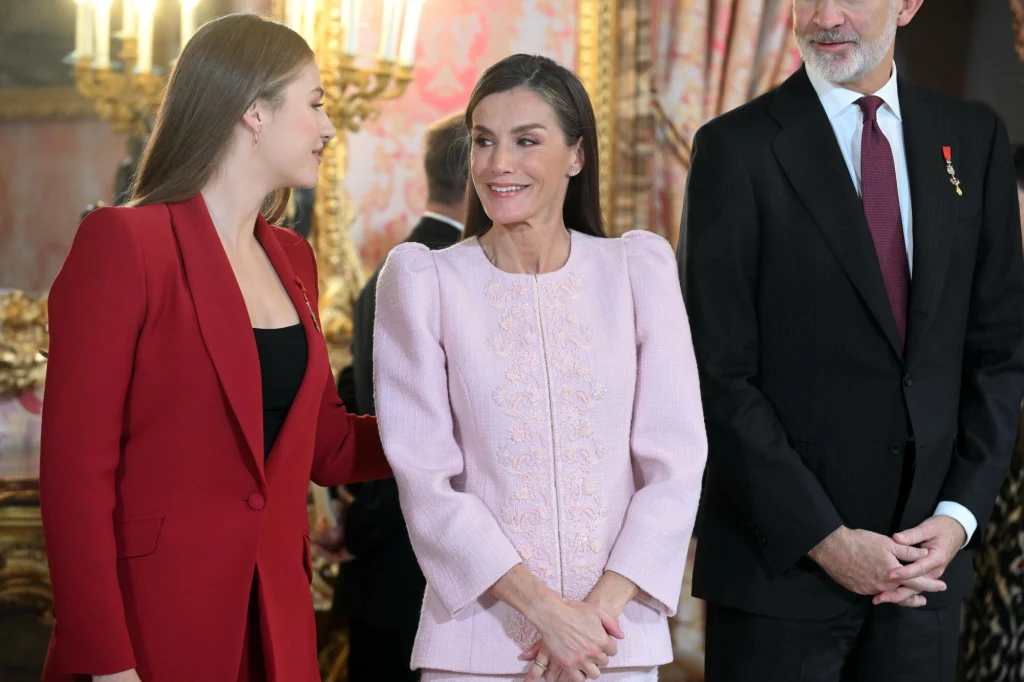 Królowa Letizia zaskoczyła wyborem stylizacji na wyjątkową okazję Trzy elegancko ubrane osoby w pomieszczeniu z bogato zdobionymi ścianami i lampami, centralna postać uśmiecha się i utrzymuje kontakt wzrokowy z kobietą w czerwonym stroju, obok mężczyzna w garniturze z przypiętym medalem.