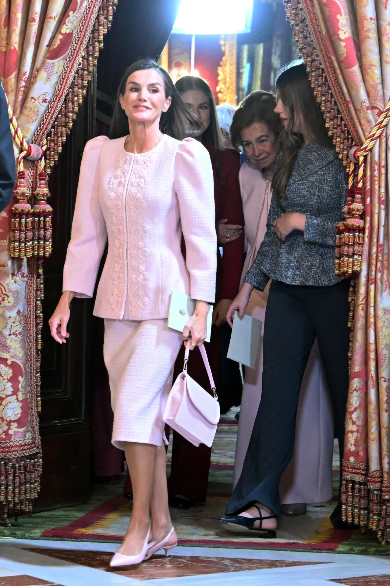 Różowy kolor i mocny krój marynarki mogły zostać uznane za dość kontrowersyjne Elegancko ubrana kobieta w jasnoróżowym kostiumie i z torebką wychodzi zza bogato zdobionej kotary, w tle towarzyszą jej inne kobiety. Atmosfera formalnego spotkania, wystrój wnętrza sugeruje miejsce o podniosłym charakterze.