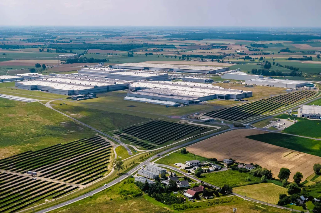 Rozległy teren przemysłowy z dużymi halami produkcyjnymi otoczonymi polami uprawnymi i infrastrukturą drogową, widoczna także instalacja paneli fotowoltaicznych oraz kilka domów w pobliżu complexu.