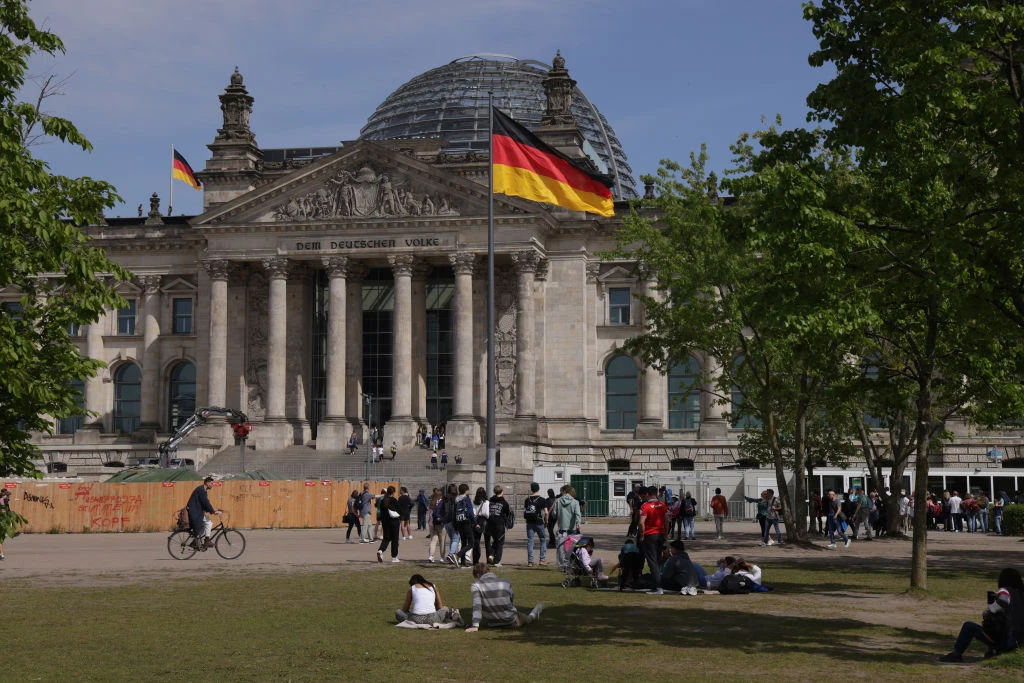 Niemiecki Reichstag z charakterystyczną szklaną kopułą, przed którym znajduje się grupa ludzi wypoczywających na trawie oraz przechodniów. Na pierwszym planie powiewa flaga Niemiec. W tle drzewa i fragmenty nieba.