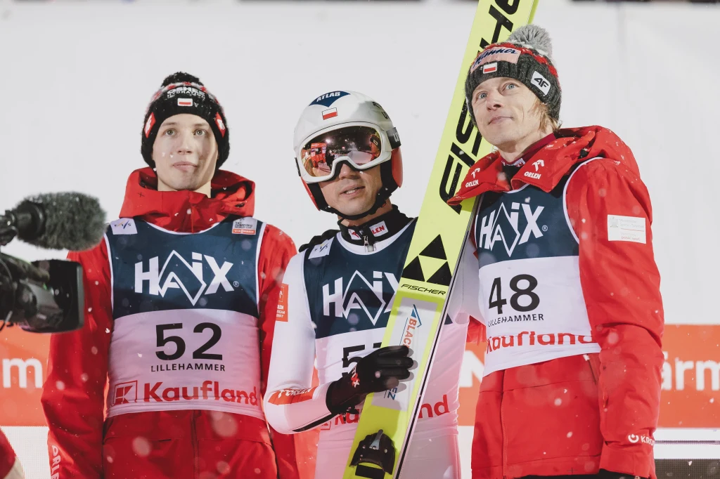 Kacper Tomasiak, Kamil Stoch i Dawid Kubacki Trzech skoczków narciarskich ubranych w czerwone kurtki i numery startowe stoi razem po zawodach, jeden z nich trzyma narty Fischer, wokół widoczny jest śnieg i elementy infrastruktury zawodów.