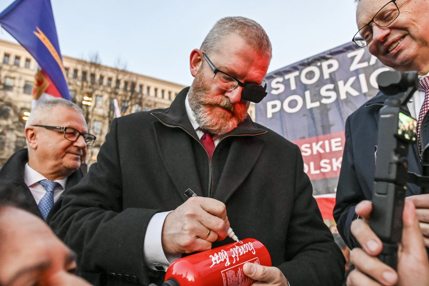 Mężczyzna z przepaską na oku podpisuje czerwoną tubę, otoczony przez inne osoby w garniturach, w tle widoczny baner z hasłami i fragment budynku.