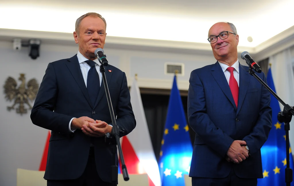 Premier Donald Tusk i nowy marszałek Sejmu Włodzimierz Czarzasty Dwóch elegancko ubranych mężczyzn stoi przy mikrofonach na tle flag Polski oraz Unii Europejskiej, obaj uśmiechnięci, jeden z nich trzyma dłonie splecione przed sobą, drugi przemawia.