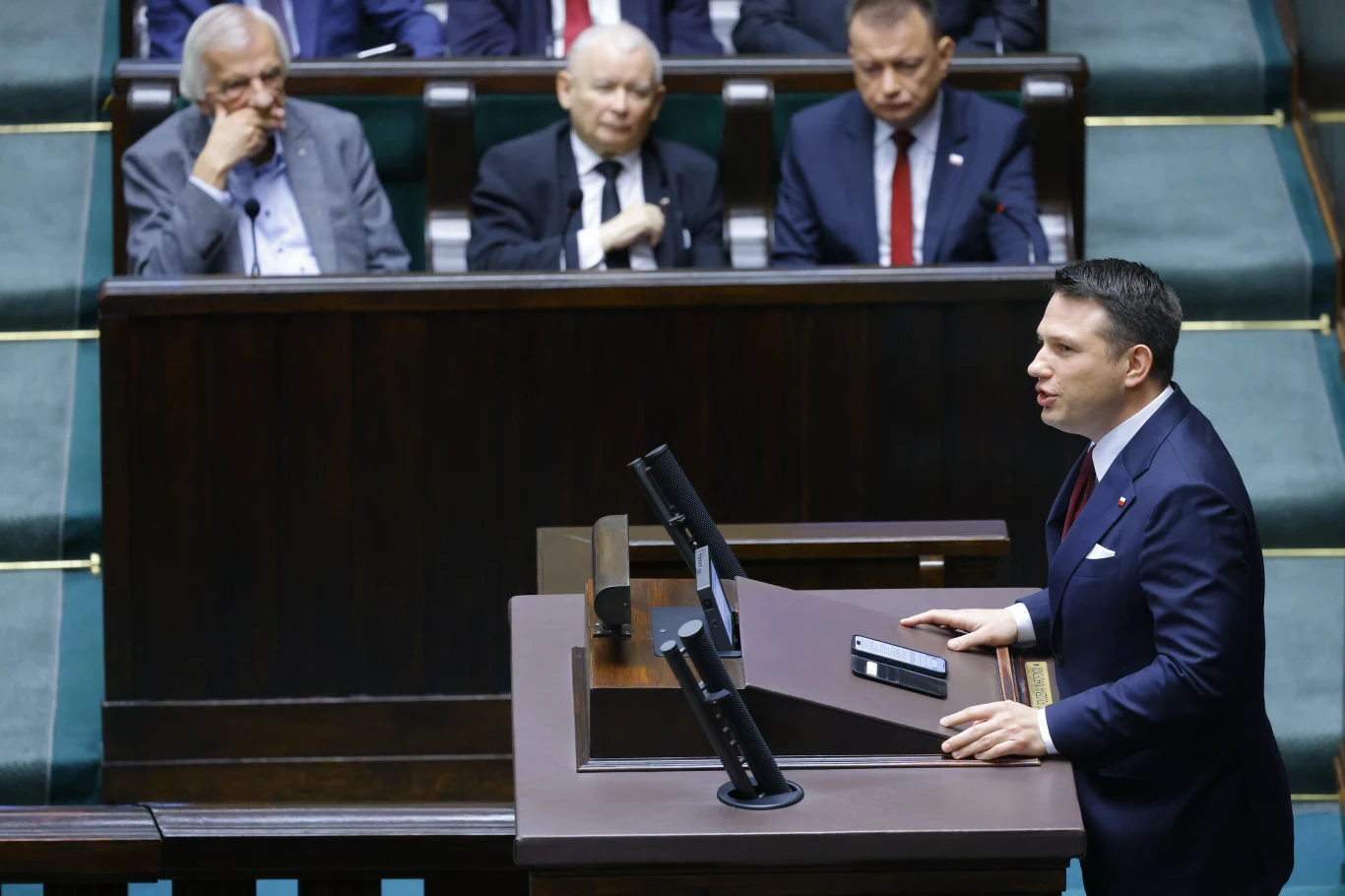 Mężczyzna w garniturze przemawia na mównicy w sali parlamentarnej, kilku polityków siedzi w ławach za nim, słuchając wystąpienia.