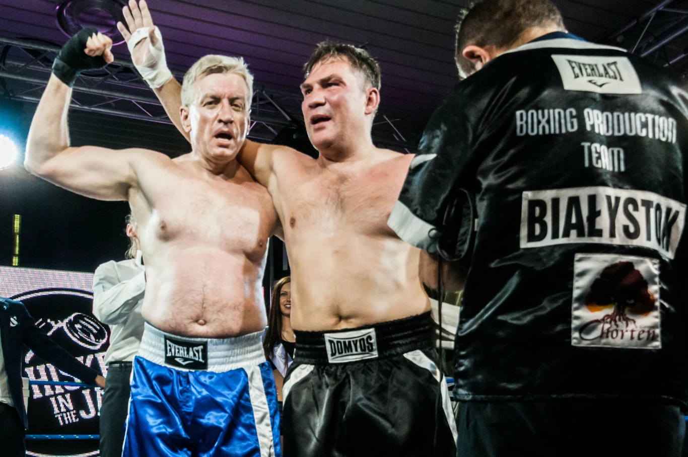 Dwóch zmęczonych bokserów stoi objętych po zakończonej walce, obaj w rękawicach bokserskich i szortach, obok nich znajduje się trener bądź członek ekipy z napisem Białystok na plecach