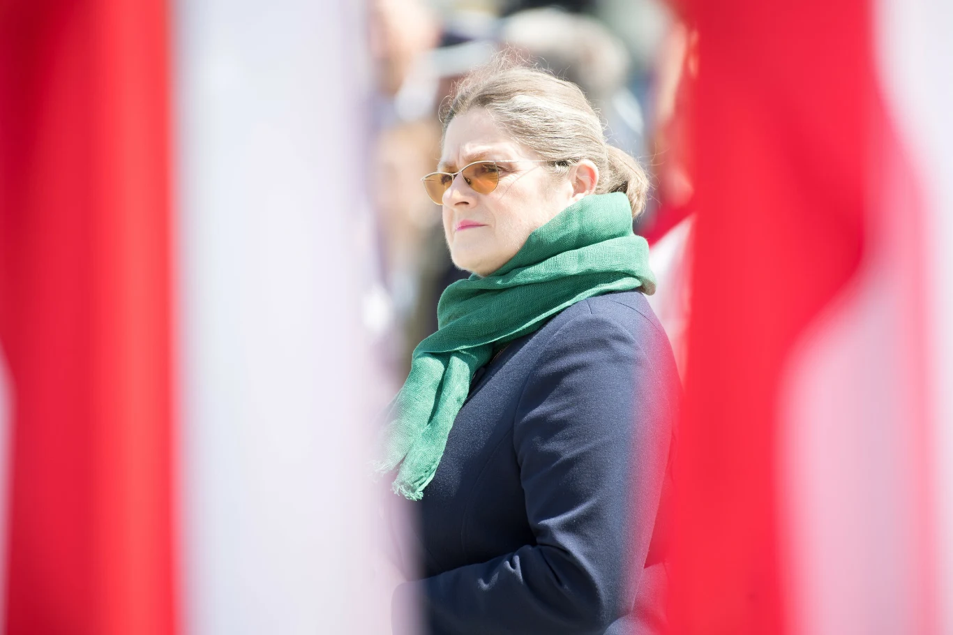 Krystyna Pawłowicz Kobieta w okularach przeciwsłonecznych i zielonym szaliku patrzy w bok, otoczona rozmytymi fragmentami dużych biało-czerwonych flag.