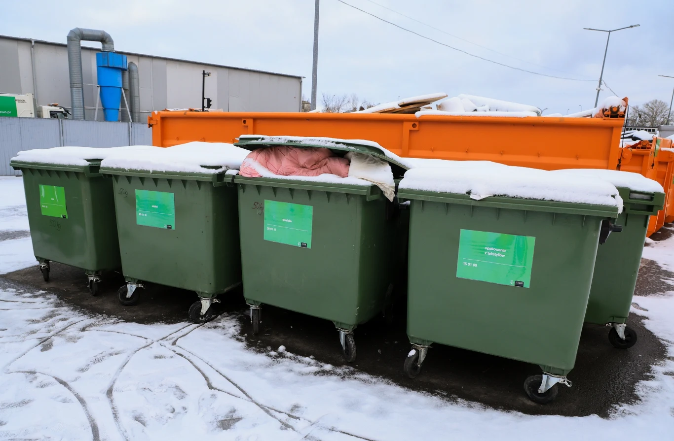 Gdzie wyrzucać stare ubrania i buty? Od 2025 roku obowiązują nowe zasady. Zielone pojemniki na odpady ustawione w rzędzie, pokryte warstwą śniegu, w tle widoczny pomarańczowy kontener oraz fragment przemysłowego budynku.