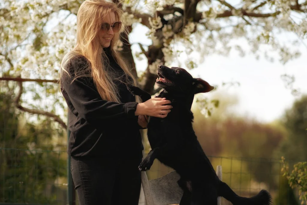 Julia Nowak stała się specjalistką w prowadzeniu psów z lękiem separacyjnym dzięki swojemu psu, Preclowi Kobieta w ciemnych ubraniach z długimi blond włosami bawi się z czarnym psem stojącym na tylnych łapach w ogrodzie na tle kwitnącego drzewa.