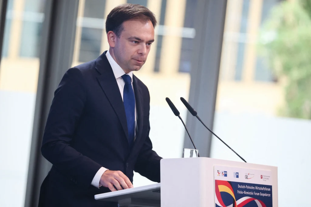 Andrzej Domański cieszy się z dobrych danych makroekonomicznych z polskiej gospodarki Mężczyzna w granatowym garniturze stojący przy mównicy podczas oficjalnego wydarzenia, przed dwoma mikrofonami, z napisem na mównicy dotyczącym polsko-niemieckiego forum gospodarczego; w tle duże okna.