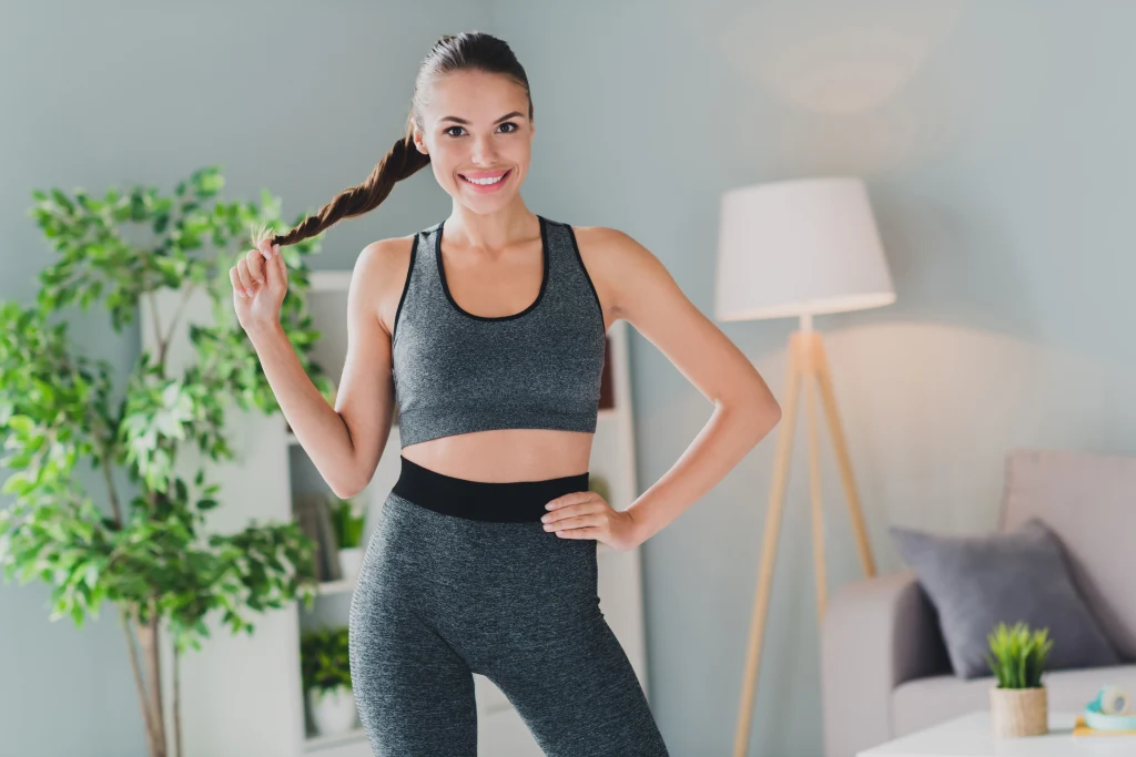 Brzuch można ćwiczyć także na stojąc Uśmiechnięta młoda kobieta w sportowym stroju, stojąca w nowocześnie urządzonym wnętrzu, trzyma się za końcówkę swojego splecionego warkocza. W tle roślina doniczkowa, lampa stojąca oraz fragment sofy.