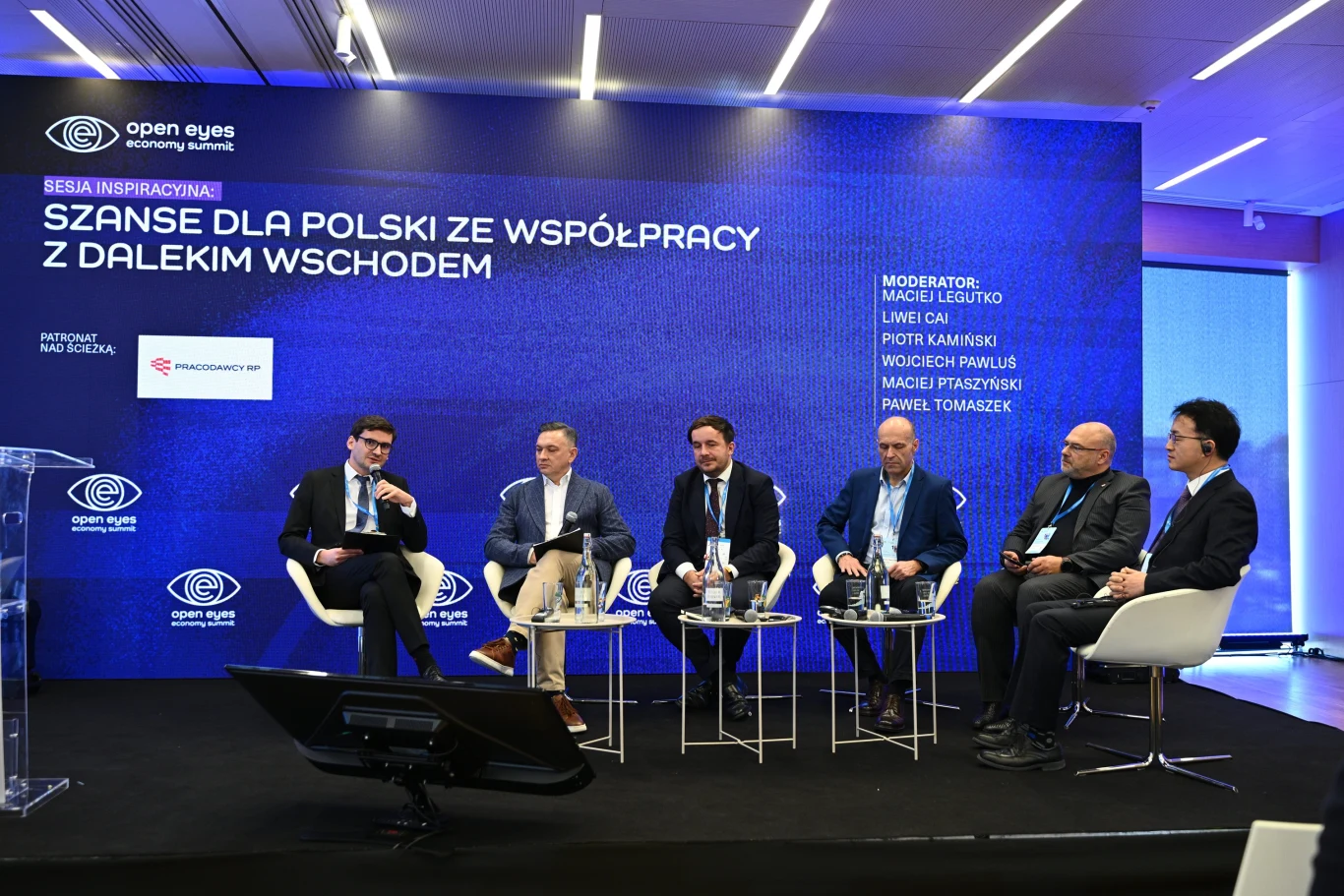 Panel dyskusyjny z udziałem sześciu mężczyzn siedzących na scenie, każdy z mikrofonem, w tle duży niebieski ekran z napisem Szanse dla Polski ze współpracy z Dalekim Wschodem oraz dodatkowymi informacjami o wydarzeniu. Sala nowoczesna, dobrze oświetlon...
