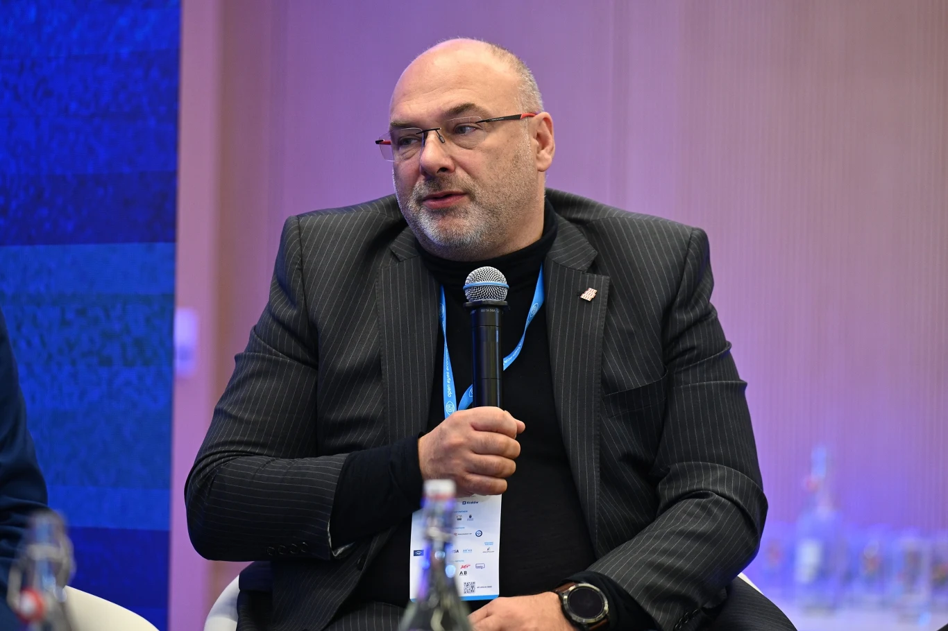 Mężczyzna w garniturze i okularach siedzi na scenie i trzyma mikrofon, za nim znajduje się tło w odcieniach niebieskiego i fioletowego, na szyi zawieszony identyfikator konferencyjny.