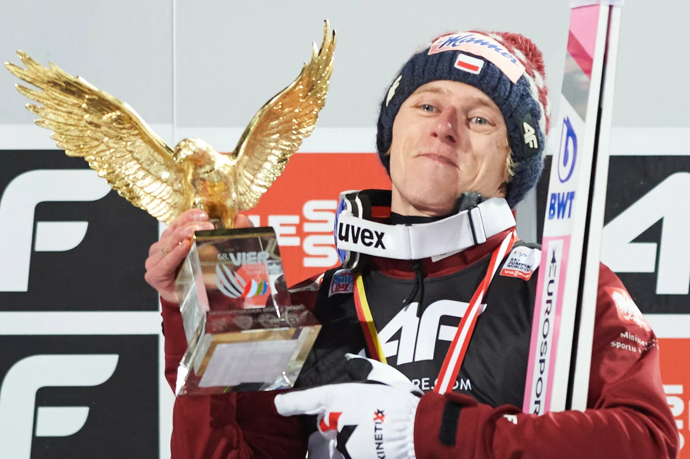 Sportowiec w kombinezonie narciarskim trzyma złotą statuetkę orła oraz narty, z medalem na szyi, na tle planszy sponsorskiej, na głowie czapka z polską flagą.