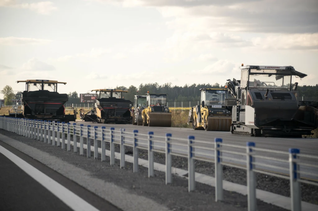 Szereg maszyn do budowy dróg ustawionych na szerokim odcinku asfaltu, po jednej stronie nowoczesna bariera drogowa, w tle las i otwarta przestrzeń.