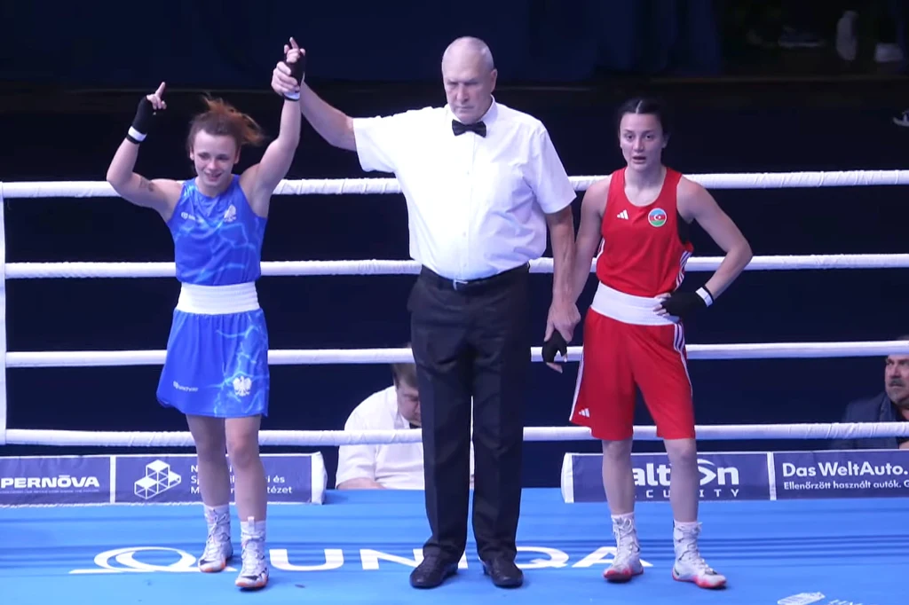 Nikola Prymaczenko (z lewej) po swojej pierwszej walce w ME U-23. Wtedy wygrała z Azerką Sędzia na ringu unosi rękę zawodniczki w niebieskim stroju, oznaczając ją jako zwyciężczynię walki bokserskiej, obok stoi zawodniczka w czerwonym stroju.