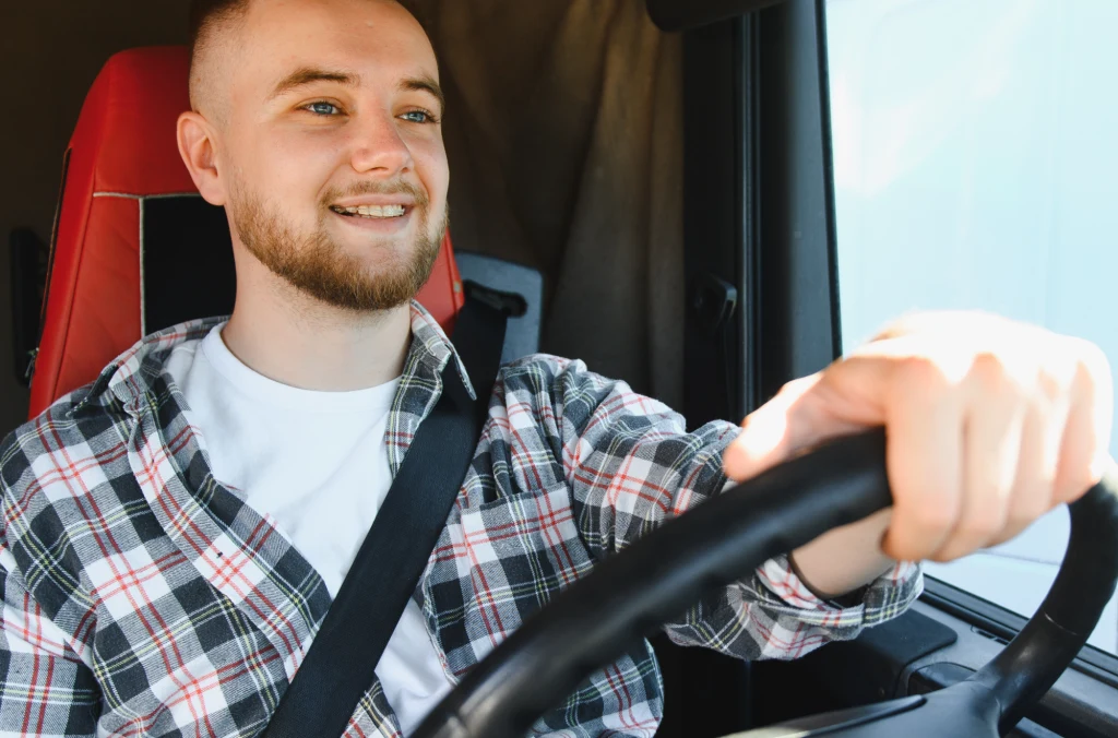 Młody mężczyzna z brodą i jasnymi włosami siedzi na czerwonym fotelu kierowcy w samochodzie, ubrany w koszulę w kratę i biały podkoszulek, uśmiecha się i prowadzi auto, trzymając kierownicę oburącz.