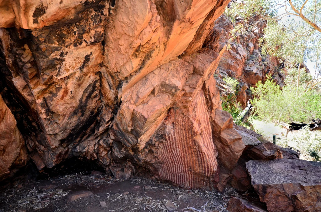 Majestatyczna czerwona formacja skalna z widocznymi warstwami geologicznymi oraz naturalnymi pęknięciami, otoczona suchą roślinnością pustynną i promieniami słonecznymi padającymi przez korony drzew.