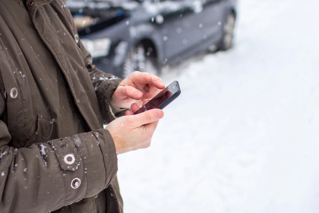 Osoba ubrana w zimową kurtkę korzysta ze smartfona na tle zaśnieżonego samochodu, otaczają ją opady śniegu.