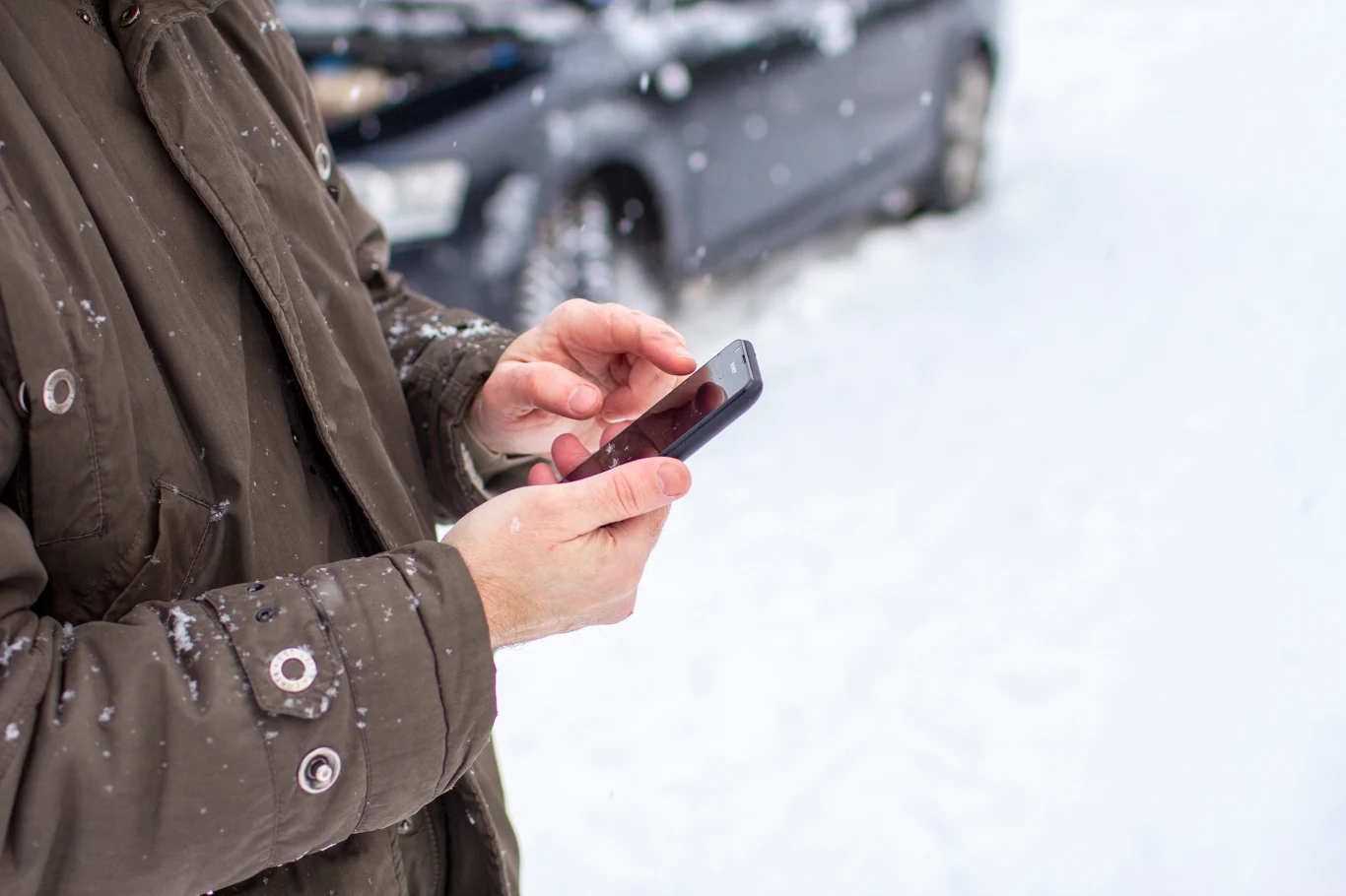 Osoba ubrana w zimową kurtkę korzysta ze smartfona na tle zaśnieżonego samochodu, otaczają ją opady śniegu.