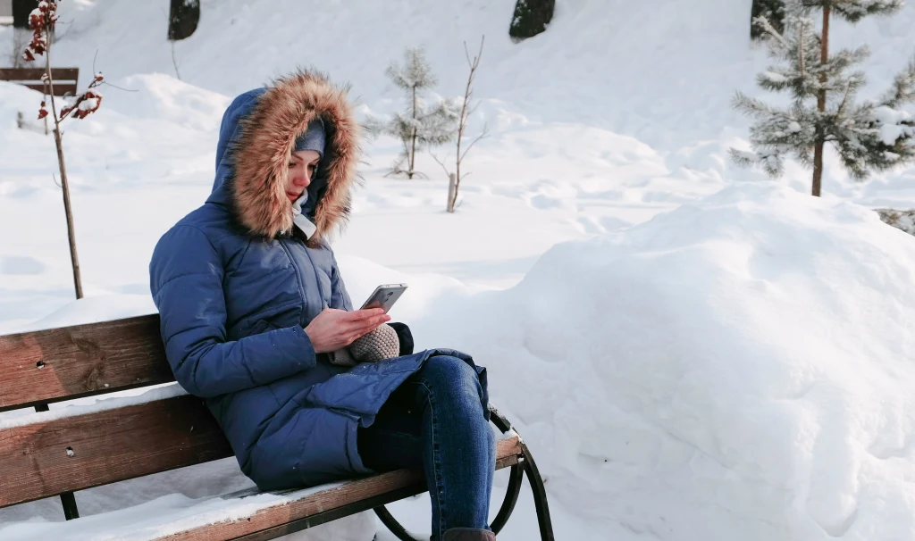 Kobieta w niebieskiej zimowej kurtce z futrzanym kapturem siedzi na ławce w parku otoczonym zaśnieżonym krajobrazem i korzysta z telefonu. W tle widoczne są drzewa i kopce śniegu.