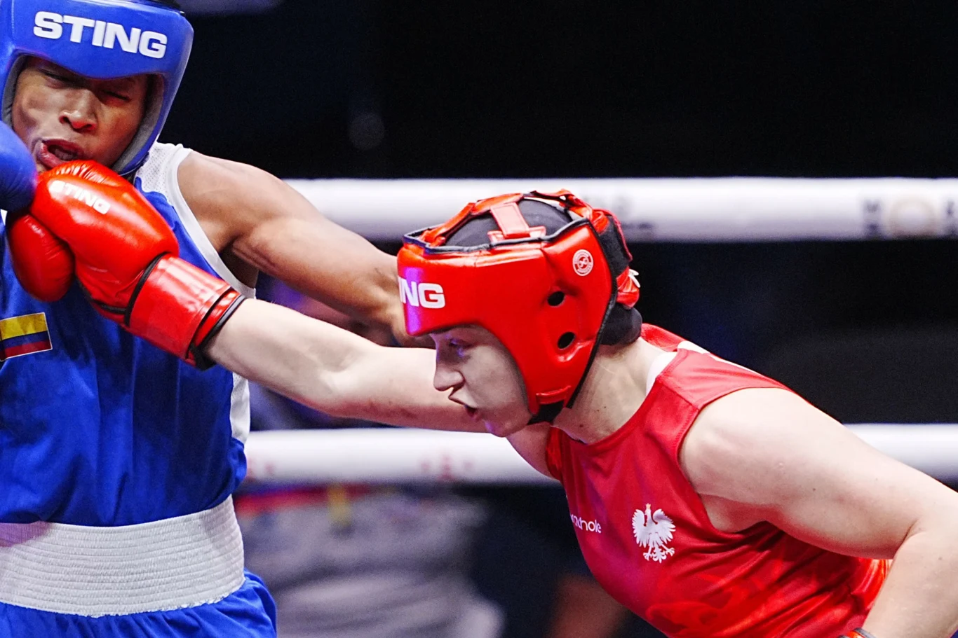Julia Szeremeta (czerwony strój) Dwóch bokserów w trakcie walki na ringu. Jeden zawodnik ubrany w niebieski strój i kask przyjmuje cios wyprowadzony przez przeciwnika w czerwonym stroju z białym orłem na piersi. Obaj sportowcy noszą kaski ochronne i rękawice bokserskie.