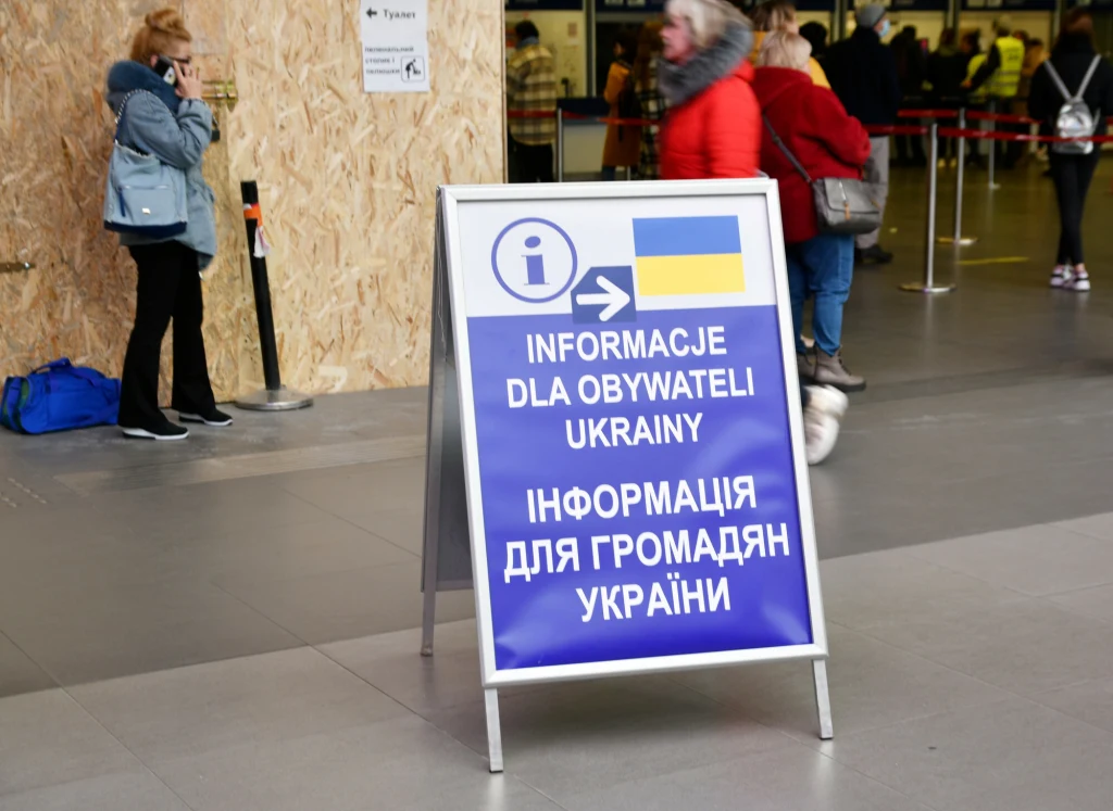 Tablica informacyjna z napisem w języku polskim i ukraińskim skierowana do obywateli Ukrainy ustawiona w przestrzeni dworca, w tle osoby oczekujące oraz ściana pokryta drewnianą płytą.