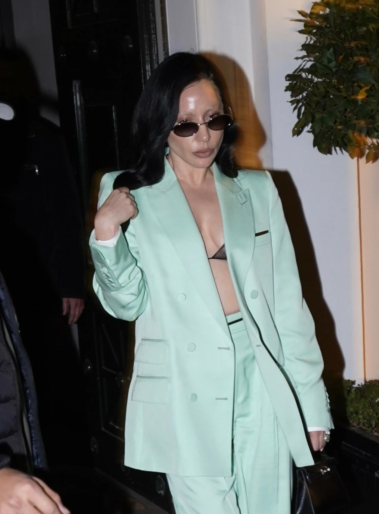 Kobieta w miętowym garniturze z rozpiętą marynarką i widocznym czarnym stanikiem, ciemne okulary przeciwsłoneczne, ciemne włosy, stoi w jasnym otoczeniu przy wejściu do budynku