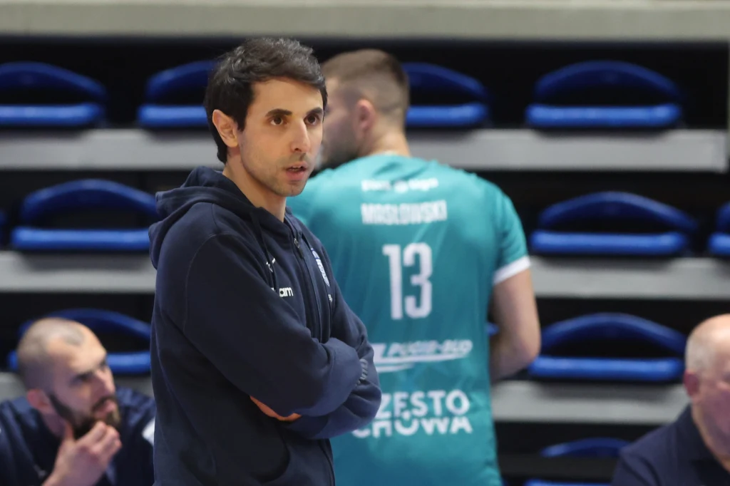 Roberto Cocconi, tymczasowy trener drużyny z Częstochowy Mężczyzna w ciemnej sportowej bluzie stoi z założonymi rękami na tle hali sportowej, w tle siatkarz w turkusowym stroju z numerem 13 oraz dwie inne osoby siedzące przy ławce rezerwowych.