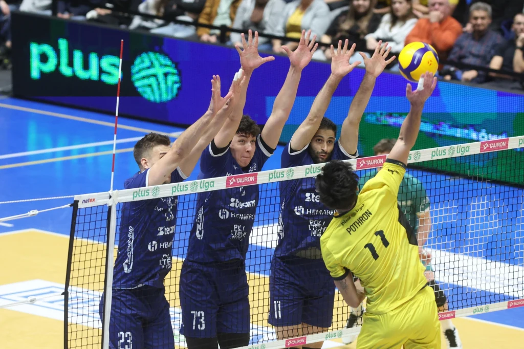 Blok siatkarzy Steam Hemarpol Politechniki Częstochowa. Drugi z lewej Jakub Nowak Trzech siatkarzy w granatowych koszulkach skacze, formując blok przy siatce, starając się zatrzymać atak zawodnika w żółtym stroju, który wykonuje zbicie piłki nad siatką podczas meczu siatkówki, a w tle widać kibiców i banery reklamowe.
