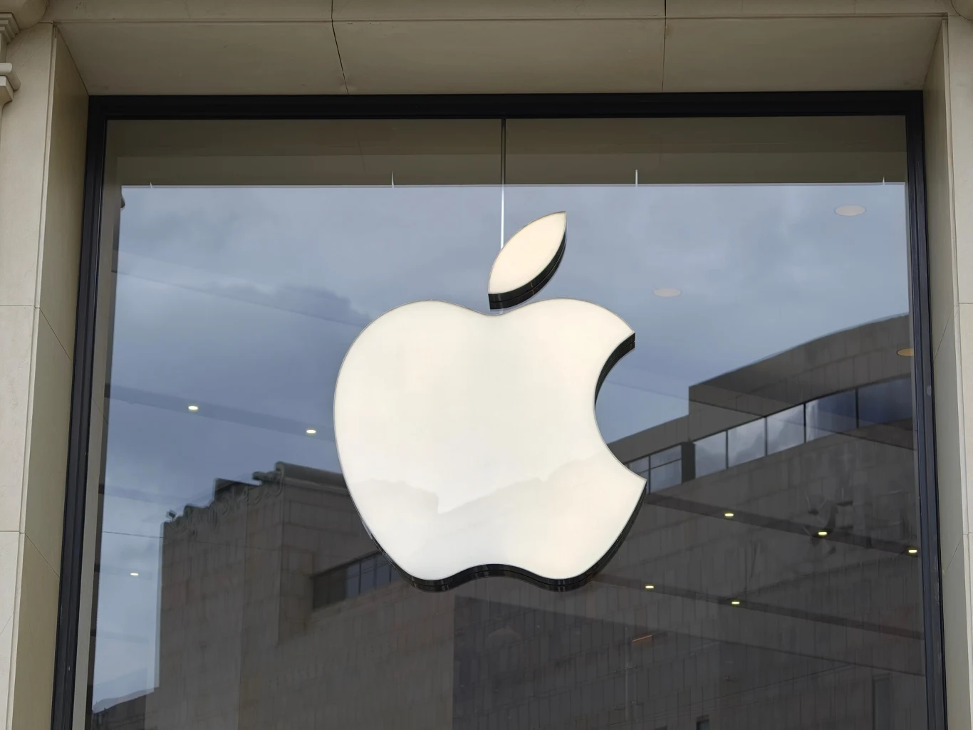 UOKiK sprawdzi działania Apple. Duże, białe logo marki Apple umieszczone na szybie witryny sklepu, w tle odbicie budynków oraz pochmurnego nieba.