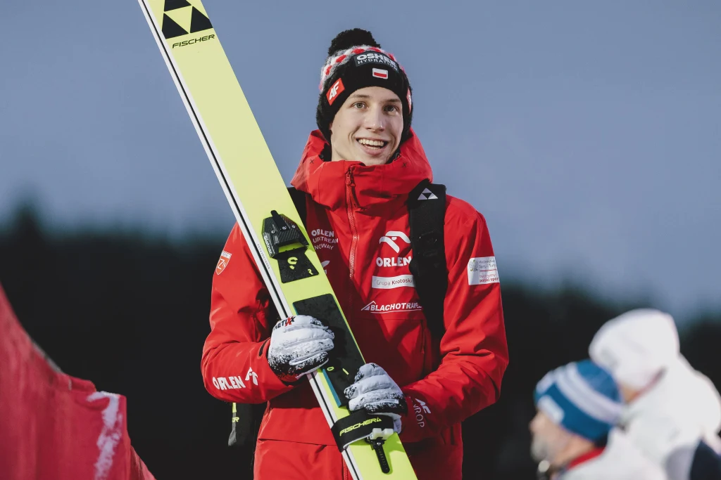 Młody mężczyzna ubrany w czerwoną kurtkę z logotypami sponsorów, czapkę zimową i rękawiczki, trzyma dużą nartę skokową i uśmiecha się, z boku znajdują się zamazane sylwetki innych osób.