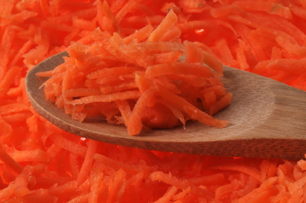 Tarta marchew świetnie nadaje się do surówek Utarta marchew na drewnianej łyżce, leżącej na tle dużej ilości startej marchewki, całość w intensywnym pomarańczowym kolorze.