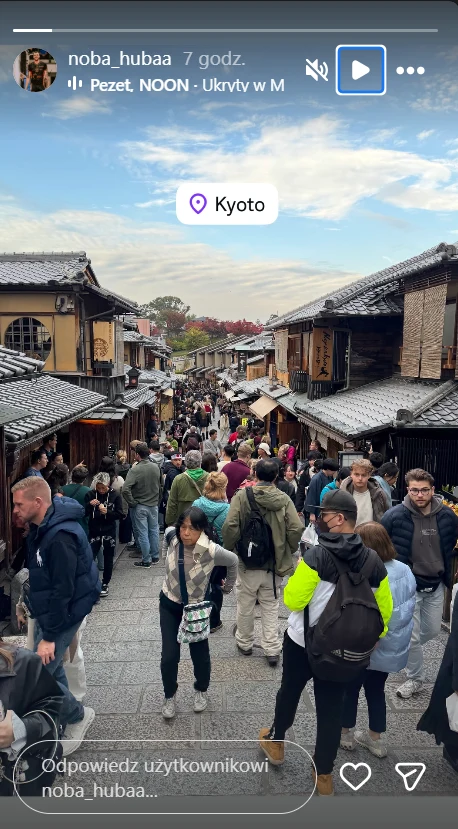 Zdjęcie Norberta Hubera Tłum ludzi spaceruje po tradycyjnej japońskiej uliczce otoczonej drewnianymi budynkami, w tle widać wzgórza i fragment nieba z lekkim zachmurzeniem.