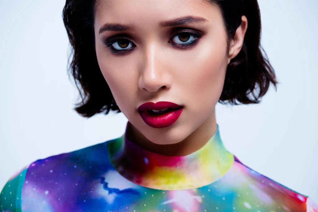 Grudniowy kolor przyciągający szczęście. Poznaj ten, który pasuje do twojego znaku zodiaku Portret młodej kobiety z ciemnymi włosami, mocno podkreślonymi oczami i ustami pomalowanymi na czerwono, ubranej w bluzkę o wzorze przypominającym galaktykę z intensywnymi kolorami.