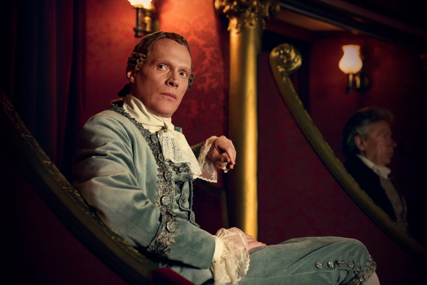 Amadeus to produkcja z udziałem laureata nagrody BAFTA® i nominowanego do Emmy® Willa Sharpe’a oraz nominowanego do nagrody BAFTA i Złotego Globu Paula Bettany’ego. Mężczyzna w stroju historycznym siedzi w loży teatralnej, elegancko ubrany w jasnoniebieski surdut z koronkowymi mankietami, na tle bordowych ścian z ozdobnym oświetleniem. Za nim rozmyta sylwetka drugiego mężczyzny, również w stylizowanym kostiumie.