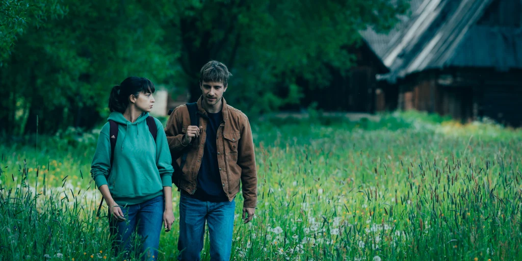 Dwie osoby spacerujące po łące otoczonej zielenią, w tle drewniane budynki wiejskie.