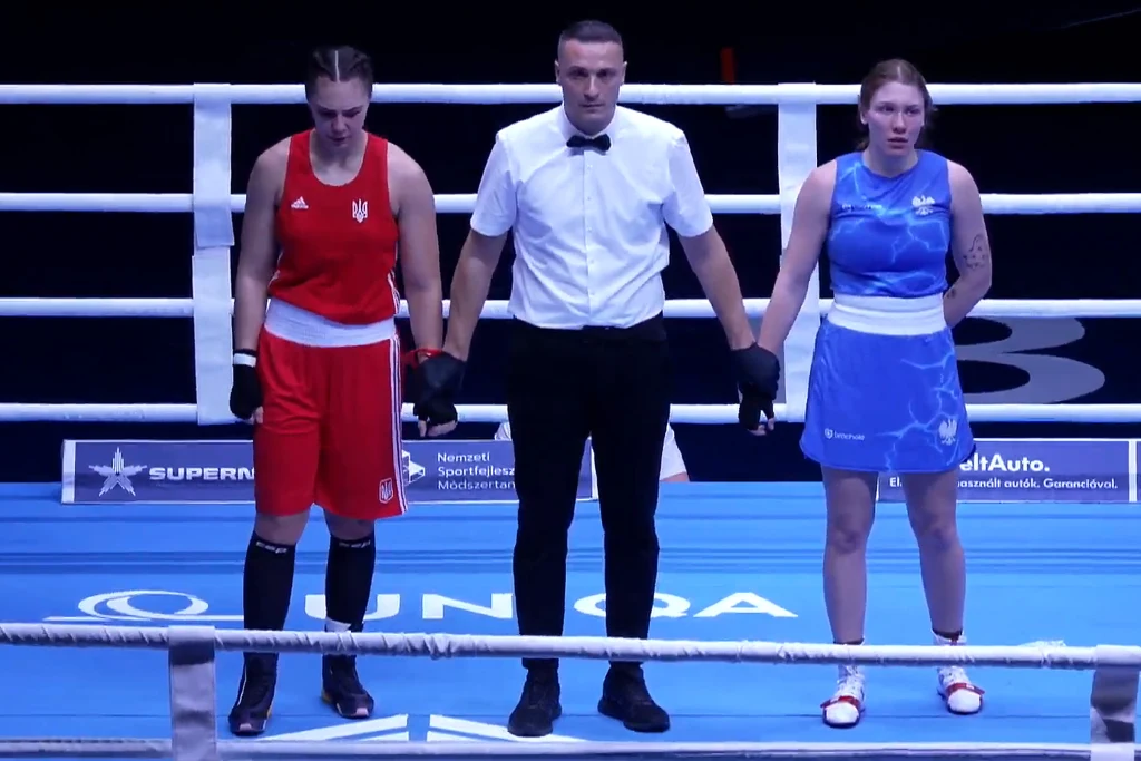 Trzy osoby stoją na ringu bokserskim, na środku sędzia w białej koszuli trzyma za ręce dwie zawodniczki, jedna ubrana w czerwony strój, druga w niebieski, obie wyglądają na zmęczone po walce.