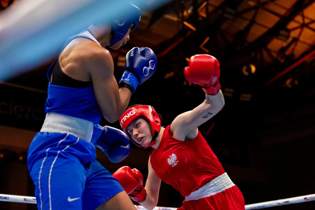 Dwie zawodniczki boksu w trakcie walki na ringu, jedna ubrana w czerwony strój z godłem Polski wykonuje cios w kierunku przeciwniczki w niebieskim stroju, obie w kaskach ochronnych, dynamiczne światło i skoncentrowanie zawodniczek podkreślają sportową ...