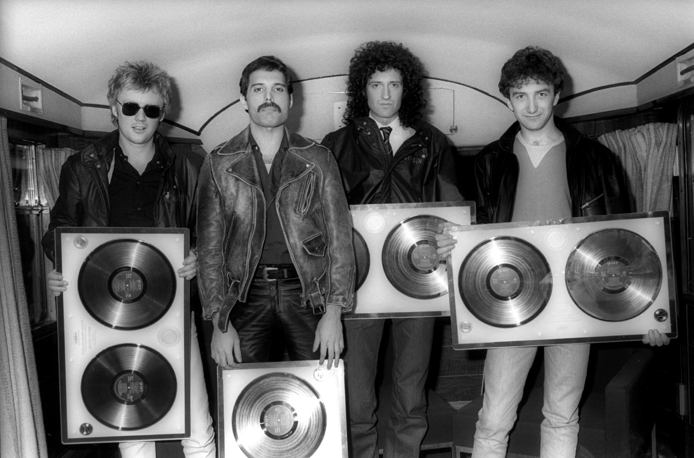 Queen w 1982 r. - od lewej: Roger Taylor, Freddie Mercury, Brian May i John Deacon Czterech mężczyzn w skórzanych kurtkach trzymających duże płyty winylowe w ramach. Tło stanowi wnętrze pojazdu lub przedziału, zasłony po bokach i uśmiechy na twarzach wskazują na celebrację osiągnięcia.