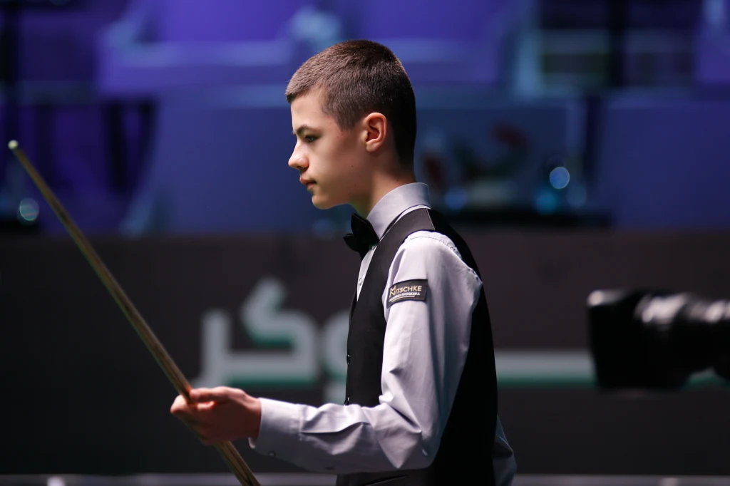 Młody chłopak w eleganckim stroju sportowym trzyma kij bilardowy, skupiony, przygotowany do wykonania kolejnego ruchu podczas profesjonalnego turnieju bilardowego.