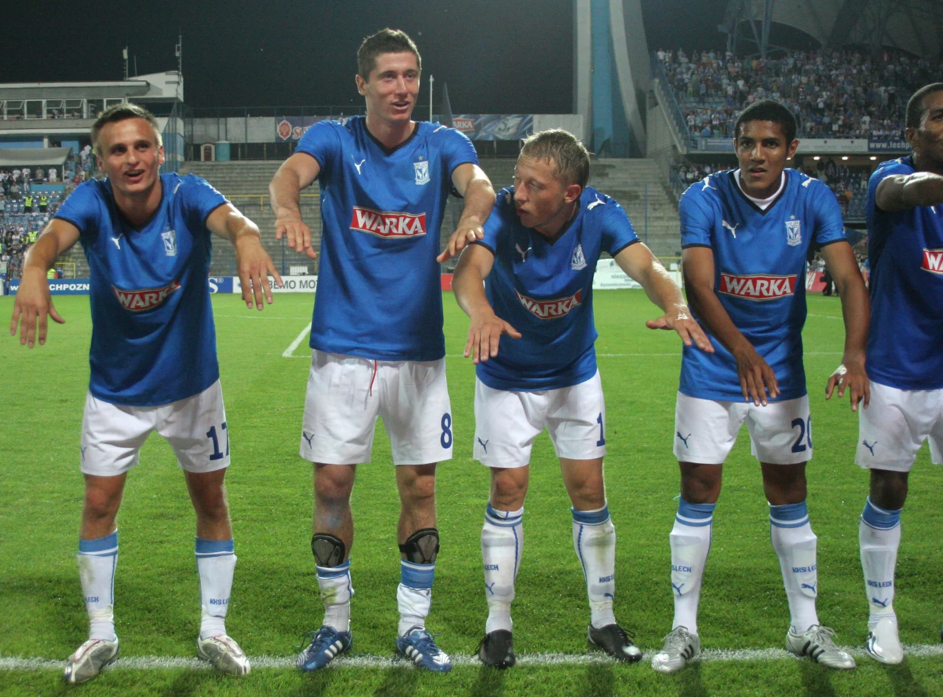 Grupa piłkarzy uśmiecha się i wykonuje humorystyczny gest rękami podczas meczu na stadionie, wszyscy ubrani w niebieskie stroje drużynowe z napisem 'Warka' i białe spodenki, w tle widoczni kibice na trybunach.